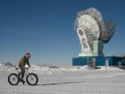 A Race Around the World participant on a bicycle with the South Pole Telescope in the background